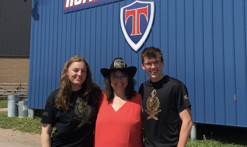 A teacher in a cowboy hat, flanked by two student, all smiling, standing in front of a building with TRHS Titans logo.