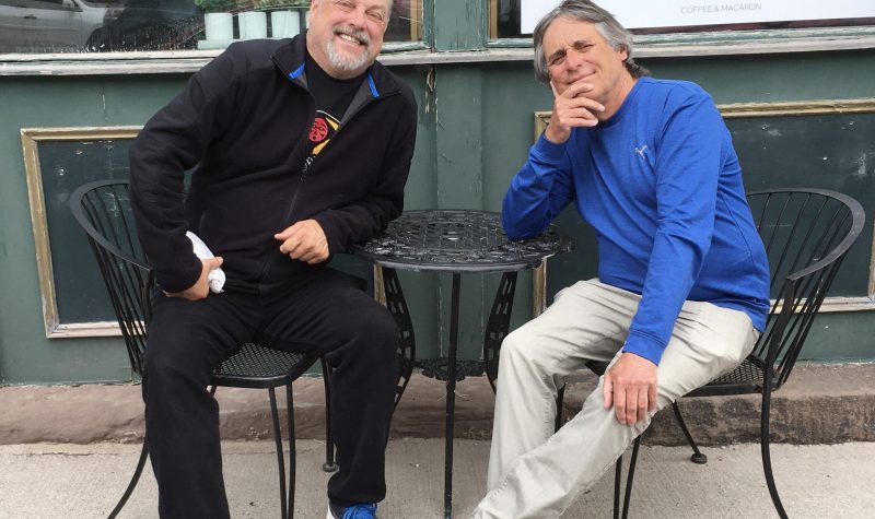 Two men sitting outside at a cafe, smiling for the camera.