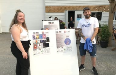 Deux personnes de l'organisation à l'extérieur avec un tableau entre les deux qui présente les affiches du festival