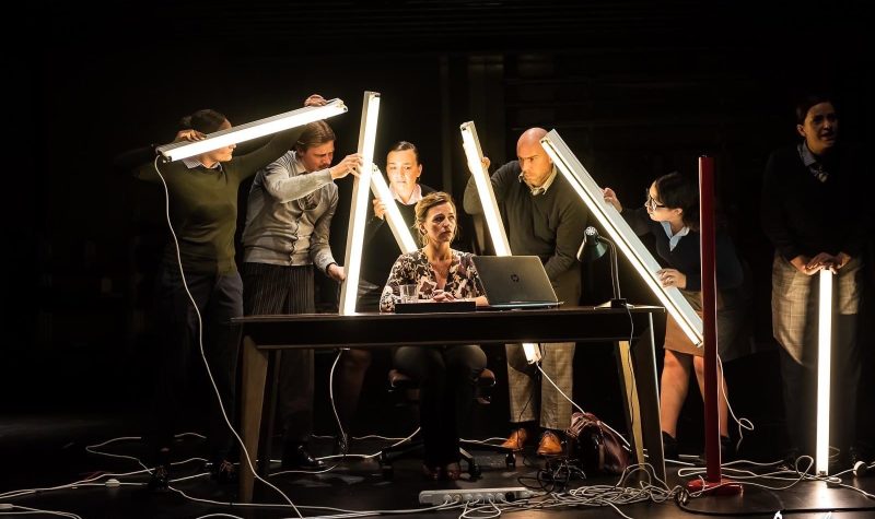 Performers hold lights sticks as they surround an actor sitting at a desk