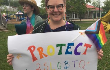 A person with short brown hair and glasses holds up a multicoloured sign saying 'Protect 2SLGBTQ+ Kids'