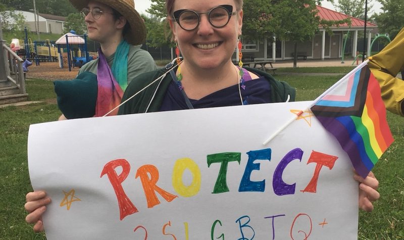 A person with short brown hair and glasses holds up a multicoloured sign saying 'Protect 2SLGBTQ+ Kids'