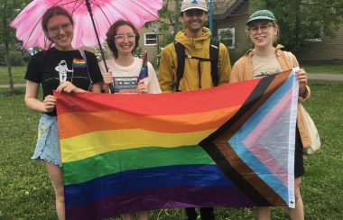Four people stand on a lawn holding the LGBT Progress Pride flag