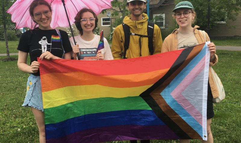 Four people stand on a lawn holding the LGBT Progress Pride flag