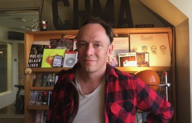 A man stands with a shelf full of zines and call letters for radio station CHMA.