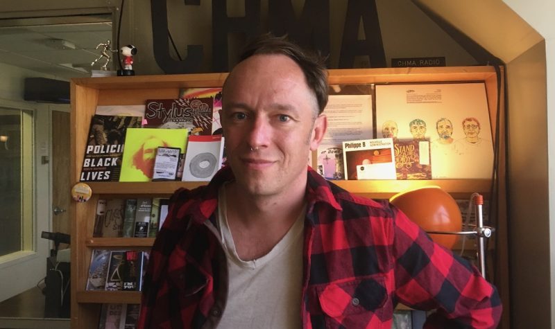 A man stands with a shelf full of zines and call letters for radio station CHMA.