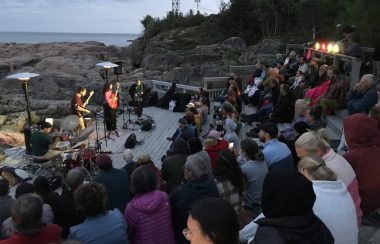 Quatre musiciens sur scène devant le fleuve Saint-Laurent entouré d'un public assis en demi-cercle