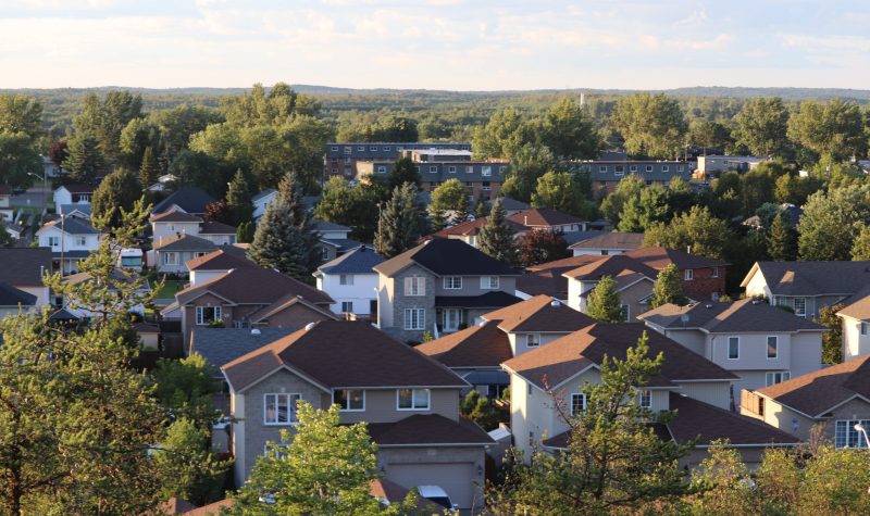 A residential neighbourhood is seen from a high angle. Many of the houses are square with brown roofs and beige siding. They are partially obscured by coniferous trees. A cloudy skyline can be seen at the top of the photo.