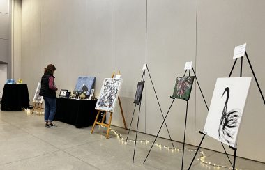A row of paintings of different animals lines a wall with a small rope of lights in the behind them. In the distance a woman looks at a table with a large painting of a bald eagle, looking at the price tag.