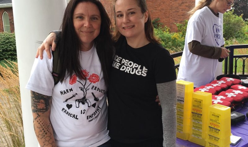 Two women stand arm in arm with naloxone kits in background