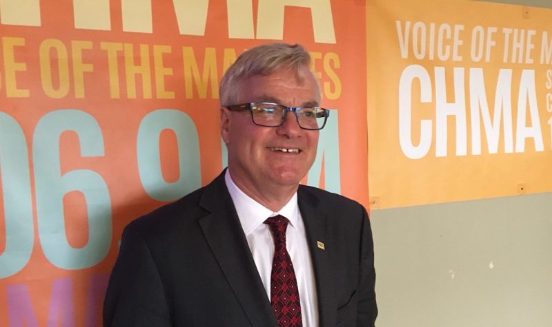 A man with suit and tie and glasses standing against a wall that reads 'CHMA Voice of the Marshes'