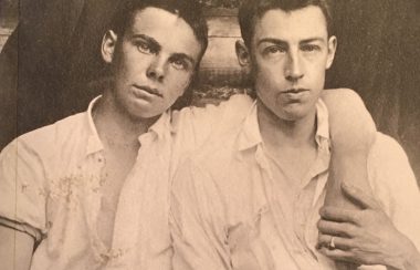Old time photo of two men, one with arm around the other, holding hands.