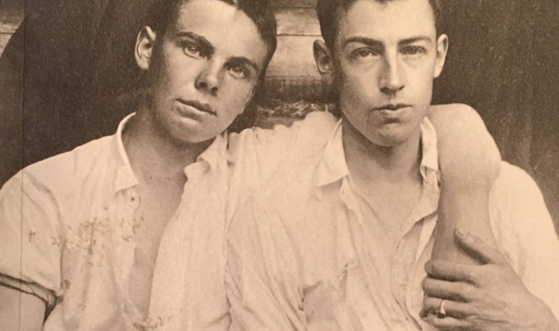 Old time photo of two men, one with arm around the other, holding hands.