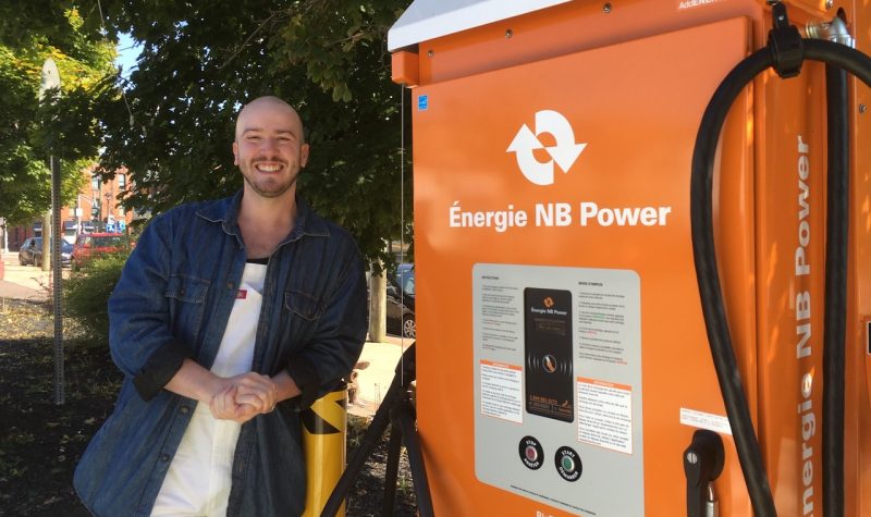 A man, smiling, beside a large orange machine with NB Power logo and thick electrical cords hanging off it.