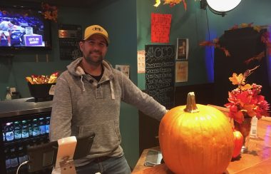 A man in a grey hoodie and brown baseball cap stands behind a bar with a pumpkin on it.