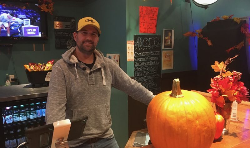 A man in a grey hoodie and brown baseball cap stands behind a bar with a pumpkin on it.