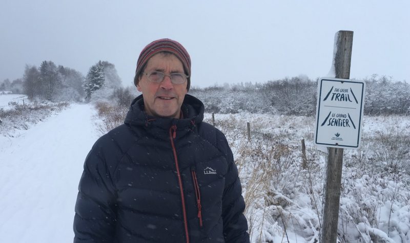 A man dressed in a black coat and tuque stands on a snowy trail next to a grassy field and fencerow. Next to him is a sign post which reads The Great Trail