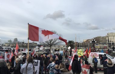 foule de personnes au bord d'une route avec des drapeau du Canada, des pancartes avec slogan et un véhicule, en arrière-plan les bateaux du port