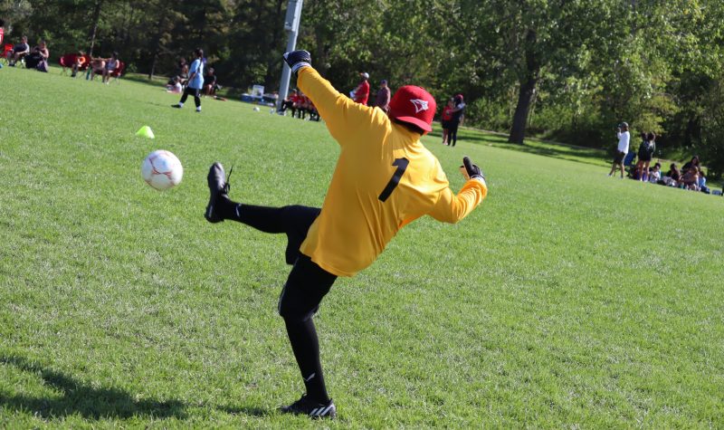 A soccer player in a yellow jersey is seen mid-kick, kicking a ball down the field to their team. Weather is sunny.