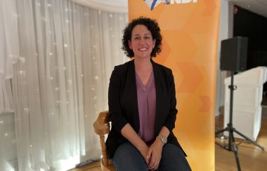 Claudia Chender is sitting smiling for a picture with the NDP orange logo behind her.