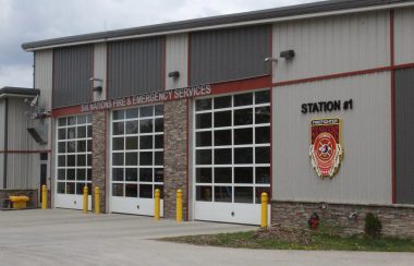 A large grey building's front entrance with 3 large garage doors with windows on each door. Above the garage doors sits a red header with the text 