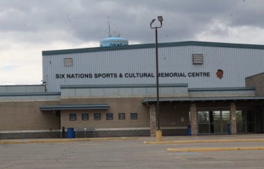 The front entrance of a large building. The top half of the exterior is a light blue colour with black writing across. The bottom half of the exterior is a light grey brick with a glass sliding door on the right side. A Light blue water tower can be seen in the distance over the building.