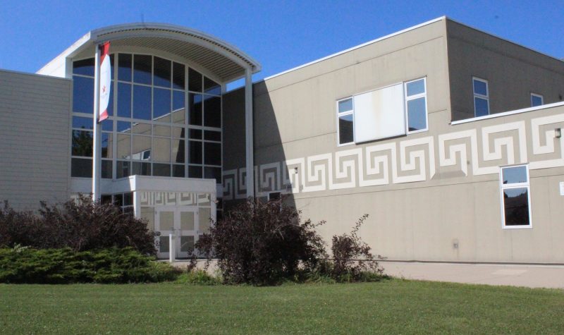 a view of a large building's front entrance. A green lawn is shown with a garden in front of the building. large glass windows can be seen extending to the top of the building along it's entrance.