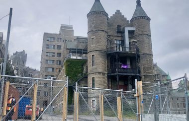 A large stone, turreted building looms behind fencing. A small Indigenous flag can be seen hanging from its side.