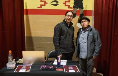 Patrick Lightning, along with one of his colleagues, stands behind a table with broachers and merch from the Maskwacis Employment Centre at a conference. The photo was taken inside a gymnasium, where the conference was taking place.