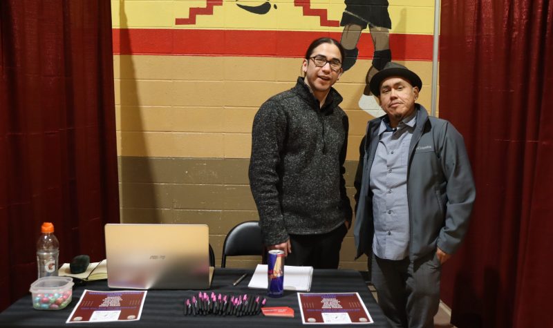 Patrick Lightning, along with one of his colleagues, stands behind a table with broachers and merch from the Maskwacis Employment Centre at a conference. The photo was taken inside a gymnasium, where the conference was taking place.