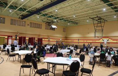 A gymnasium full of chairs and roundtables as people eat lunch and listen to the speakers at the front of the room. There are also water agencies with booths in the back of the gym.