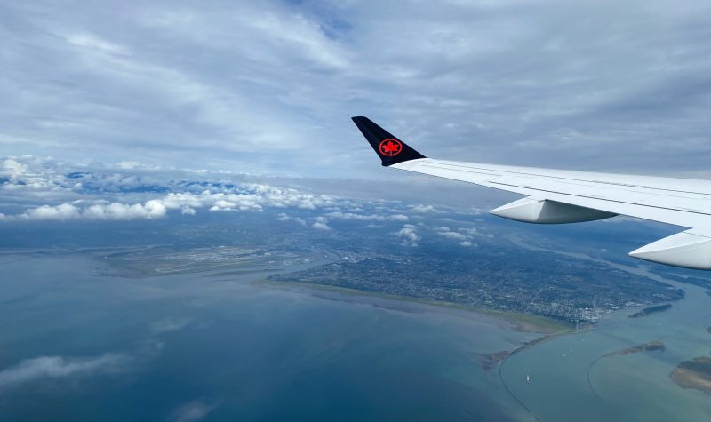 Vue aérienne de l'océan et d'une ville côtière, sous des nuages blancs, captée du hublot d'un avion, vis-à-vis de l'aile.