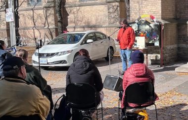 A person in a red jacket is speaking into a microphone to a seated crowd. They are outside infront of a brick building.