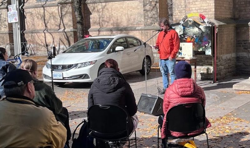 A person in a red jacket is speaking into a microphone to a seated crowd. They are outside infront of a brick building.