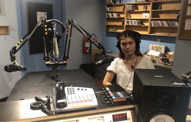 A young woman sits in front of a mic in a radio recording studio. Equipment is in the foreground and shelves of CDs in the background.