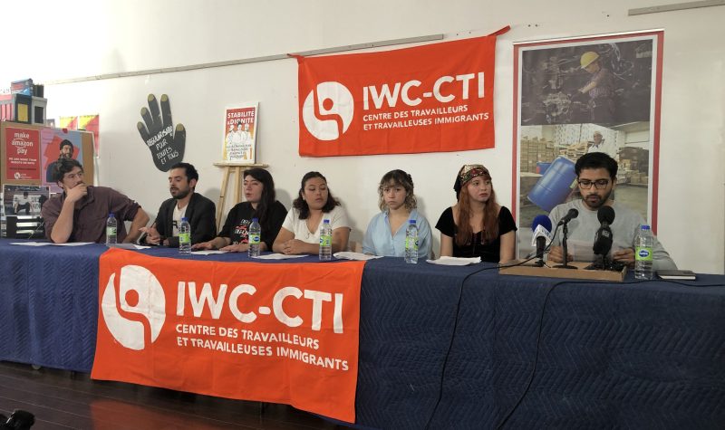 A group of people sit behind a table at a press conference, with Immigrant Workers Centre banners in front and behind them.