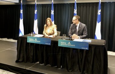 A woman and a man sit at a table with Quebec flags behind them.