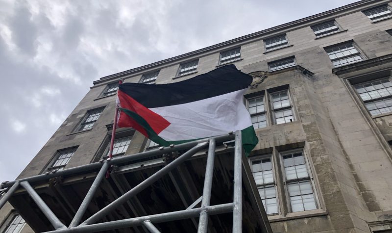 A Palestinian flag flies atop scaffolding in front of a building entrance. A stormy sky can be seem in the background.