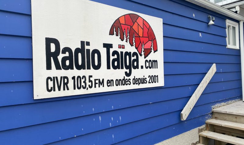 Sur le mur de bois bleu d'une maison, à gauche des escaliers, l'affiche de Radio Taiga CIVR 103,5 FM