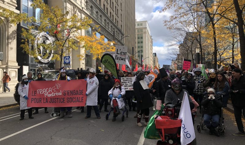 A group of hundreds, many in costume, gathers on a city street in autumn. They are holding protest banners and union flags.