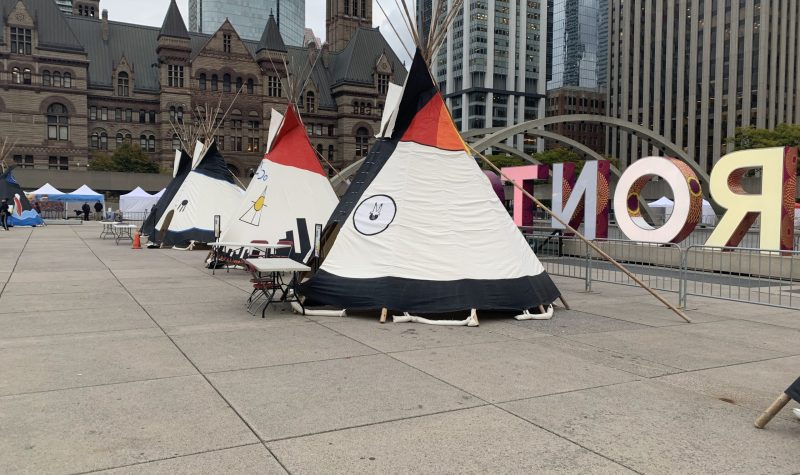 Tipis of different colours outside with a sign and building behind them.