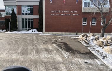 Bâtisse du campus Saint-Jean. La bâtisse est de couleur brune. Il y a les drapeux du Canada, de l'Alberta et de la Francophonie albertaine accroché à l'entrée. Il y est inscrit Faculté Saint-Jean sur le mur d'entrée.