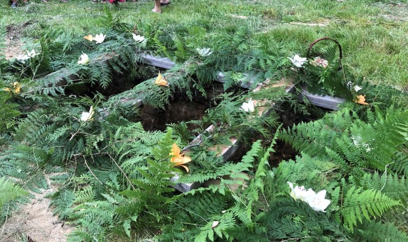 Ferns and flowers are arranged on top of wooden boards at the surface level of as yet unfilled grave.