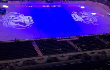 La patinoire du Rogers Place. Les lumières sont éteintes dans l'aréna. Des projections de deux logos des Oil Kings se retrouvent dans chaque zone de la patinoire. Quelques lumières bleues et rouges sont projetées sur la patinoire.