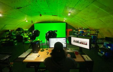 Un homme de dos, cheveux longs, est assis devant deux écran, face à des projecteurs qui pointes verts un grand écran vert.