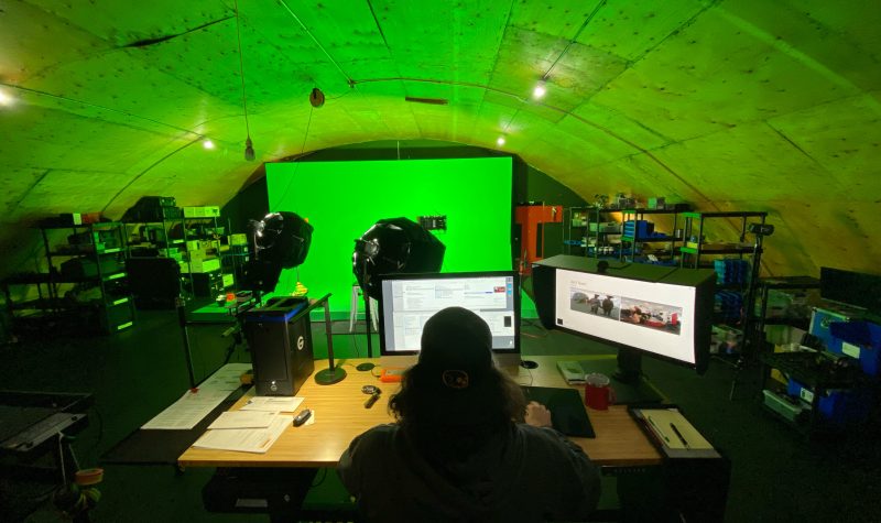Un homme de dos, cheveux longs, est assis devant deux écran, face à des projecteurs qui pointes verts un grand écran vert.