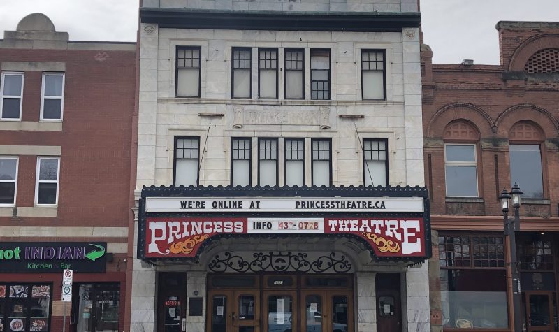 Entrée du cinéma Princess. La bâtisse est blanche et elle est haute de 3 étages. L'enseigne Princess Theatre est inscrit au-dessus de l'entrée du cinéma en blanc et rouge.