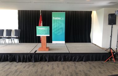Salle ayant un podium d'annoncement avec un pupitre au centre. Des chaises se trouvent à la gauche du pupitre d'annoncement. À droite, on aperçoit un haut-parleurs. Une affiche PrairiesCan en turquoise est placé derrière le pupitre d'annoncement avec le drapeau du Canada.