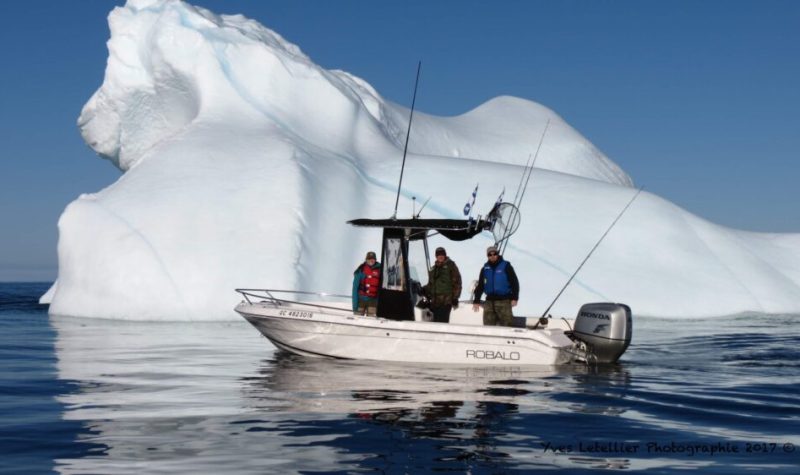 Un bateau sur l'eau avec trois personnes à son bord, devant un iceberg trois fois sa taille.