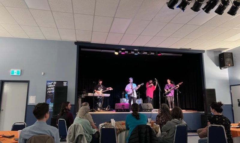 Sept personnes sont assises à des tables rondes devant une scène. Sur scène, quatre personnes jouent de la guitare, du banjo et du clavier devant des micros.